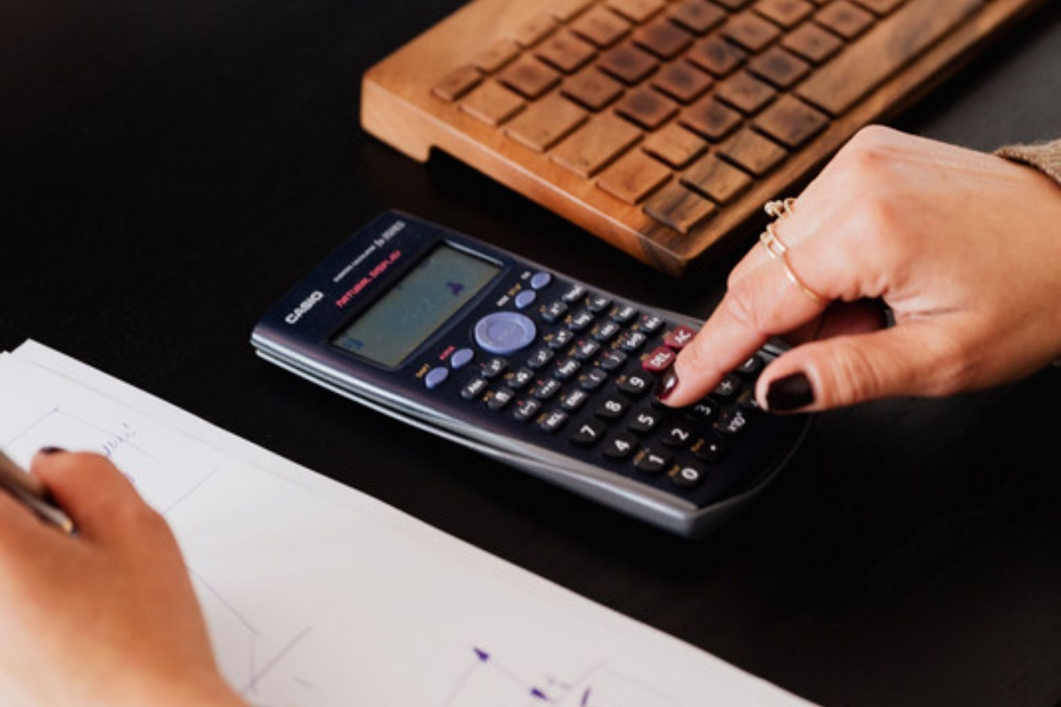 Hånd taster på en lommeregner ved et tastatur, mens den anden hånd skriver noter