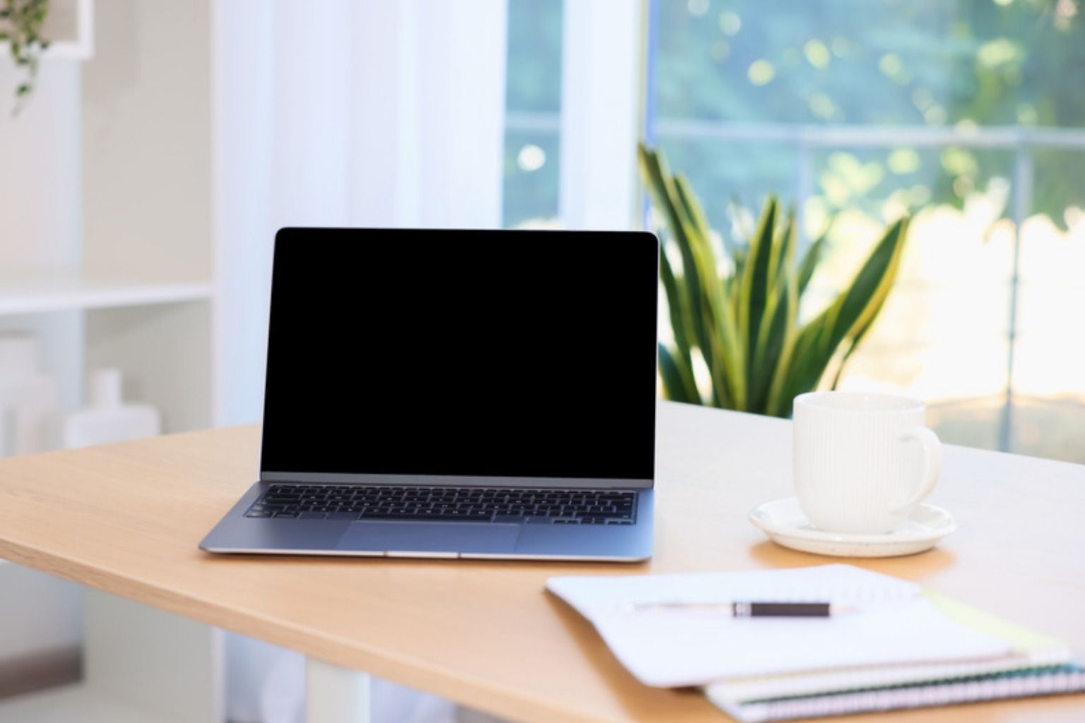 Bærbar computer, kaffekop og notesbog på et lyst skrivebord ved et vindue